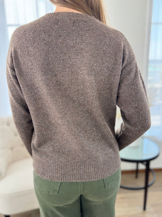 A woman viewed from behind wearing a heathered taupe-brown knit sweater and olive green pants in a bright indoor room.