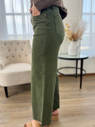 A person wearing olive green wide-leg pants, shown from the waist down in a bright indoor room with a tufted chair, side table, and wood floor.