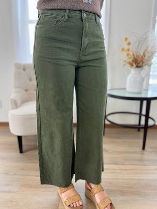 A person wearing olive green wide-leg cropped pants with side slit details, paired with woven slide sandals in a bright indoor room with a chair and side table in the background.