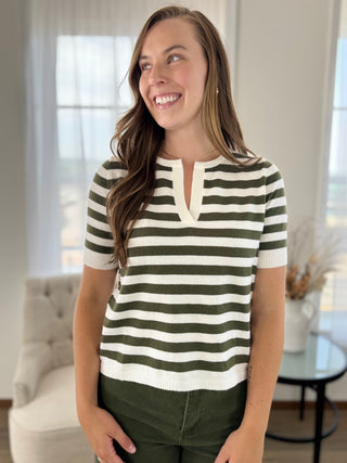 A woman wearing a short-sleeve knit top with horizontal olive green and off-white stripes, paired with olive green pants, standing in a bright, modern living room.