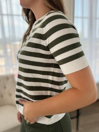 A woman wearing a short-sleeve knit top with horizontal olive green and off-white stripes, shown from the side in a softly lit indoor setting.