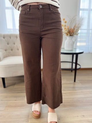 A person wearing brown wide-leg cropped pants with a striped top and cream platform sandals, standing in a bright living room with a tufted chair and side table in the background.