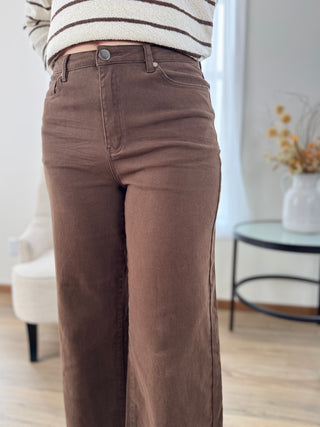 A person wearing brown wide-leg corduroy pants and a cream and brown striped sweater, standing in a bright, modern room with a white chair and a small round table in the background.