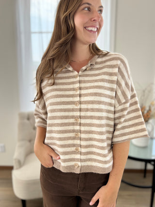 A woman wearing a beige and cream horizontally striped short-sleeve knit button-up cardigan, paired with brown pants, standing in a brightly lit indoor room.