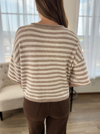A woman viewed from behind wearing a beige and brown horizontally striped knit top with short wide sleeves, paired with brown pants, standing in a brightly lit room with sheer curtains.