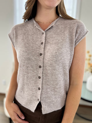 A woman wearing a light taupe heathered knit button-front cardigan vest with cap sleeves, paired with dark brown pants.