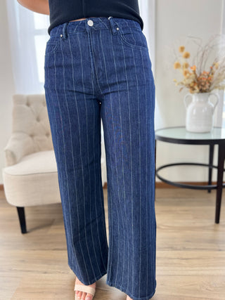 A person wearing dark blue wide-leg pinstripe jeans, standing in a bright living room with a tufted chair and a side table in the background.
