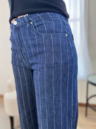 Close-up of a person wearing blue denim jeans with thin white vertical pinstripes, paired with a dark navy top in an indoor room setting.