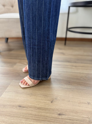 A person wearing dark blue pinstriped trousers and beige slide sandals, standing on a light wood laminate floor in a home setting.
