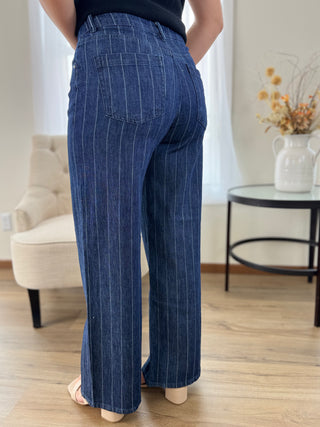 A person shown from the back wearing dark blue wide-leg pinstripe jeans in a bright indoor room with a chair, side table, and vase of dried flowers.