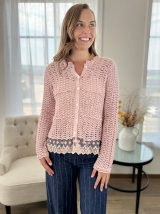 A woman wearing a light pink hand-crocheted button-up cardigan with a gray lace hem, paired with dark blue pinstriped jeans, standing in a bright, modern room.