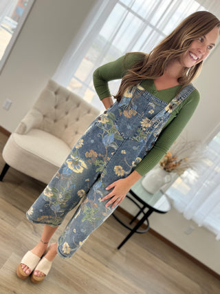 A woman wearing a blue floral print overall jumpsuit over an olive green long-sleeve top, standing in a bright living room.