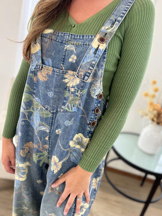 A person wearing blue floral denim overalls over a green ribbed long-sleeve top, shown from the shoulders to the thighs in an indoor room setting.