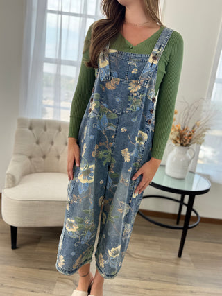 A woman wearing a green ribbed long-sleeve top and blue floral denim overall jumpsuit, standing in a bright living room with a tufted armchair and a small round table.