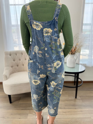 A person viewed from behind wearing blue denim overall-style shorts with a large cream and green floral print, layered over a ribbed olive green long-sleeve top in a bright indoor room with a chair, side table, and window.
