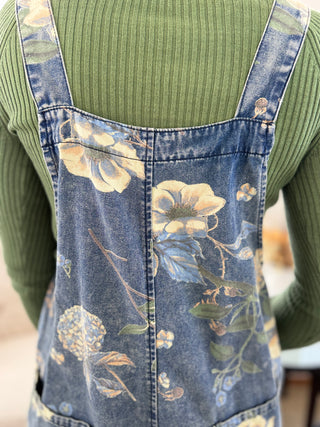 A close-up of a person wearing blue denim overalls with a faded floral print over a ribbed sage green long-sleeve top.