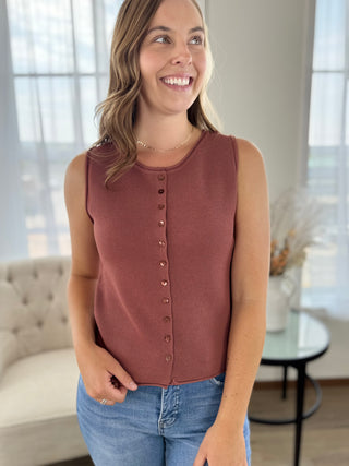 A woman wearing a sleeveless dusty rose knit top with a button-down front, paired with blue jeans, standing in a brightly lit room.