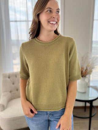 A woman wearing a short-sleeve knit sweater in a muted olive green color, paired with blue jeans, standing in a brightly lit living room.