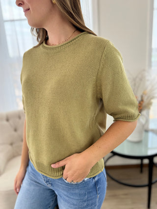 A woman wearing a short-sleeve knit sweater in a muted olive green color, paired with light blue jeans, standing in a bright, modern room.