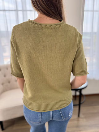 A woman viewed from behind wearing a short-sleeve ribbed knit sweater in a muted olive green color, paired with blue jeans in a bright indoor setting.