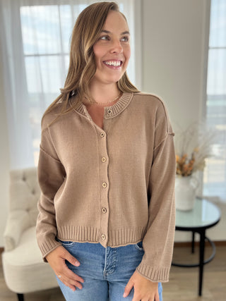 A woman smiling and wearing a cropped, long-sleeve knit cardigan in a warm taupe-beige color with matching buttons, paired with blue jeans in a bright, modern living room setting.