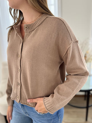 A woman wearing a taupe knit cardigan sweater with button-front closure and ribbed collar, cuffs, and hem, paired with blue jeans in a softly lit indoor setting.