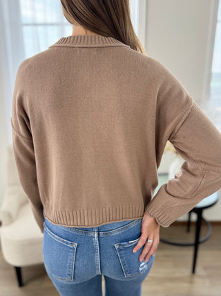 A woman viewed from behind wearing a cropped, long-sleeve knit sweater in a warm taupe-brown color, paired with blue jeans in a bright indoor setting.