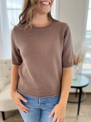 A woman wearing a short-sleeve crewneck knit sweater in a taupe brown color, paired with blue jeans, standing in a brightly lit living room.
