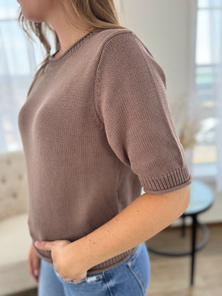 A woman wearing a short-sleeve knit sweater in a dusty mauve-brown color, paired with blue jeans, shown from a side-back angle in a bright indoor setting.