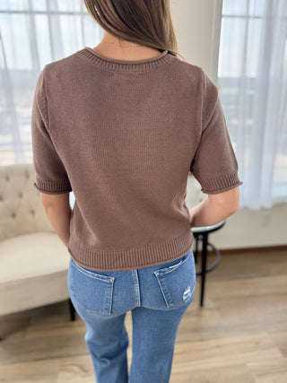 A woman viewed from behind wearing a short-sleeve ribbed knit sweater in a warm taupe-brown color, paired with blue jeans, in a brightly lit room with sheer curtains and a light-colored sofa.