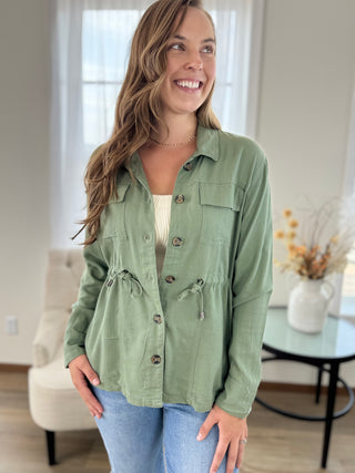 A woman wearing a sage green button-up shirt jacket over a cream top and blue jeans, standing in a bright, modern living room.