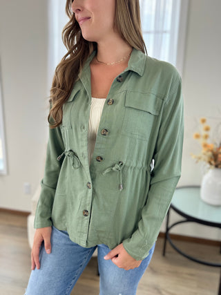 A woman wearing an olive green button-up shirt jacket with chest and waist tie details, styled over a white ribbed top and blue jeans in a bright indoor setting.