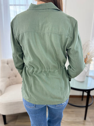 A person shown from the back wearing an olive green utility-style jacket with a drawstring waist, paired with blue jeans in a bright indoor room.