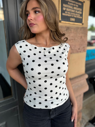 A woman wearing a white ribbed sleeveless top with black polka dots, paired with dark jeans, standing outdoors in front of a stone wall and plaque.