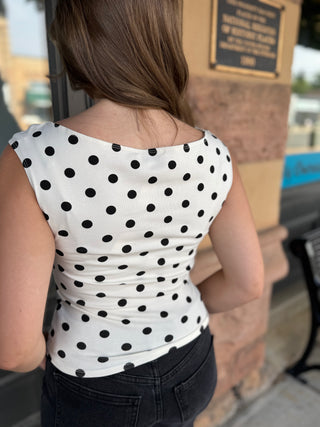 A woman viewed from behind wearing a white sleeveless top with black polka dots, paired with dark jeans, standing outdoors near a stone pillar.