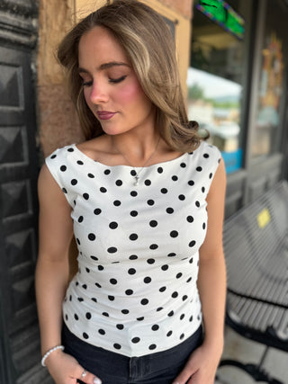 A woman wearing a white sleeveless top with black polka dots, paired with dark jeans, standing outdoors in front of a storefront.