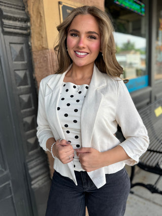 A woman wearing a white textured blazer over a black and white polka dot top, paired with dark gray jeans, standing outdoors in front of a storefront.