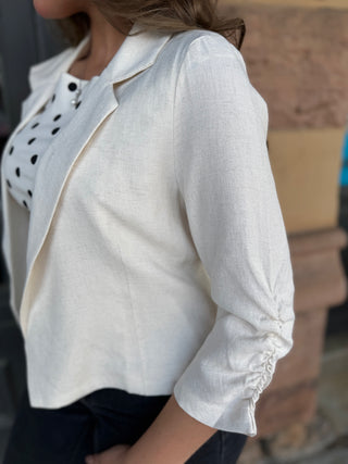 A woman wearing a white linen blazer with three-quarter sleeves and gathered cuffs, layered over a black and white polka-dot top.