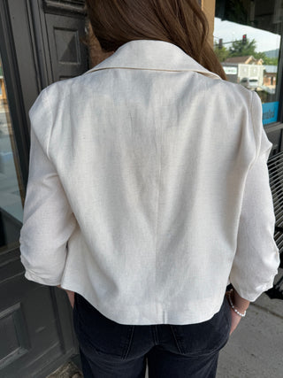 A person viewed from behind wearing a white linen-style cropped jacket with three-quarter sleeves and dark jeans, standing outdoors near a dark door.