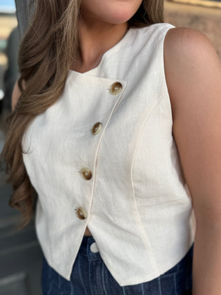 A close-up of a woman wearing a sleeveless off-white linen vest with a diagonal button closure and dark blue jeans.