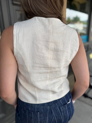 A woman viewed from behind wearing a sleeveless off-white linen top and dark blue pinstriped pants, standing in an outdoor setting.