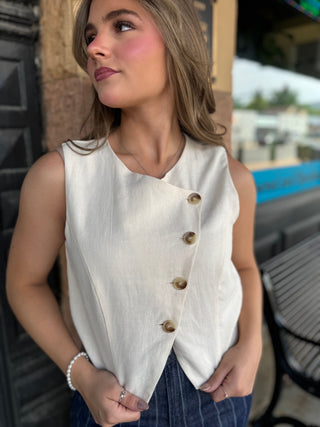 A woman wearing a sleeveless cream-colored wrap-style vest with brown buttons, paired with dark blue jeans, standing outdoors.