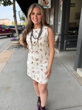 A woman stands on a sidewalk in front of a storefront, wearing a sleeveless cream dress with a cowboy-themed print including boots, horseshoes, and cactus. She is accessorized with layered brown beaded necklaces and dark purple boots.