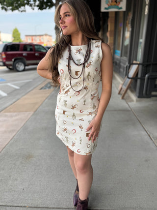 A woman wearing a sleeveless cream-colored mini dress with a brown and tan western print, accessorized with layered brown beaded necklaces, standing on a city sidewalk.
