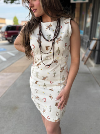 A woman wearing a sleeveless cream-colored mini dress with a western-themed print of cacti, horseshoes, and cowboys, accessorized with layered brown beaded necklaces, standing on a city sidewalk.