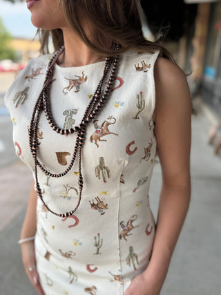 A woman wearing a sleeveless cream-colored dress with a western-themed print of cacti, horseshoes, and cowboy boots, accessorized with layered dark brown beaded necklaces.