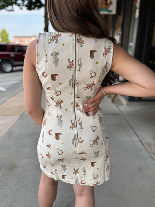 A woman viewed from behind wearing a sleeveless bodycon dress with a western-themed print of cacti, horseshoes, and tumble dogs on an off-white background. The dress features a central zipper and is photographed outdoors on a sidewalk.