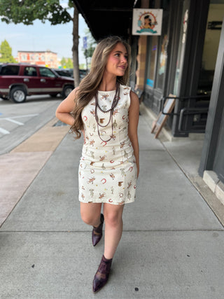 A woman walking on a city sidewalk wearing a sleeveless cream mini dress with a western-themed print of horseshoes, lasso, and boots, paired with dark brown ankle boots and layered necklaces.