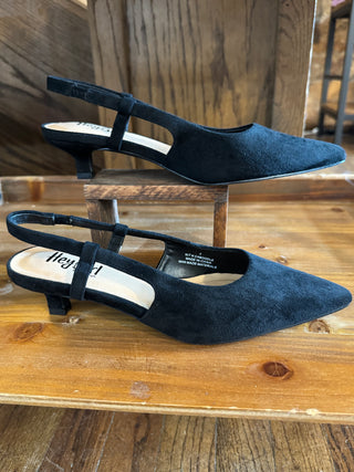 A pair of black suede slingback pointed-toe pumps with low block heels, displayed on a rustic wooden shelf with one shoe resting on a small wooden block.
