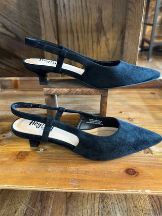 A pair of black suede slingback pointed-toe flats with low block heels, displayed on a rustic wooden surface with one shoe resting on a small wooden stand.